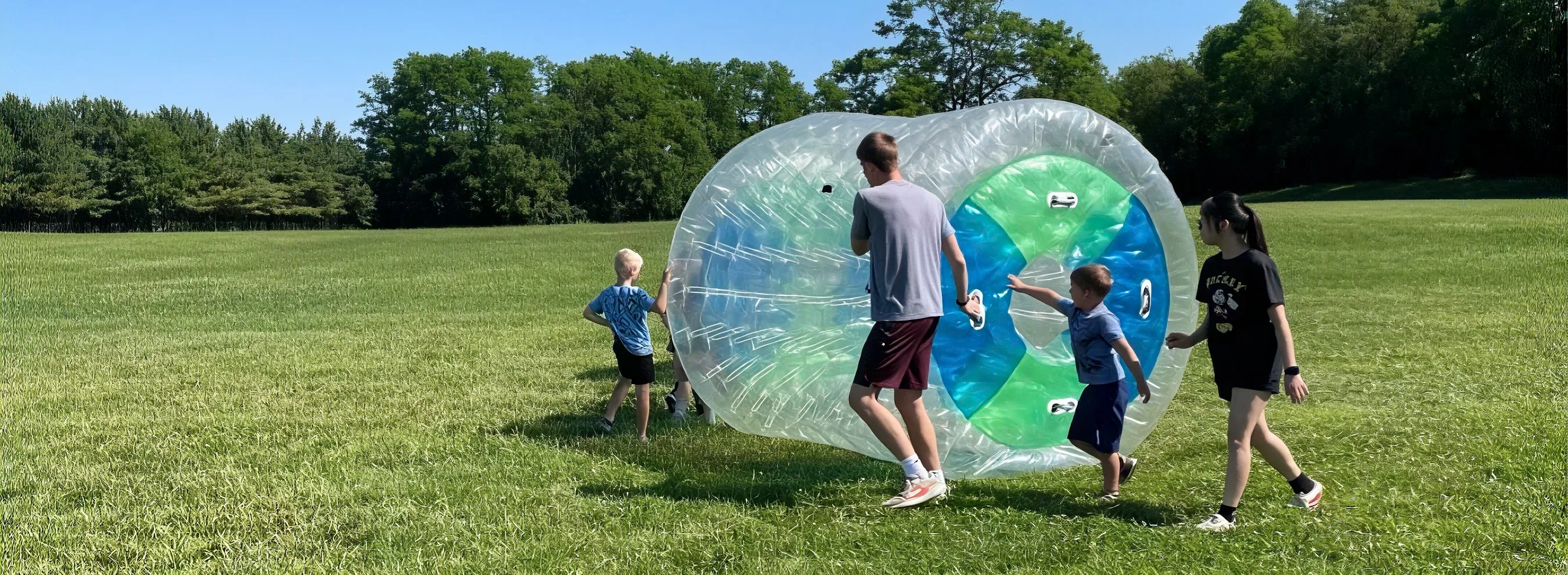 A camp counselor rolls a giant inflatable ball while two boys reach through side openings and a girl walks alongside during an outdoor game at Journey Camp.