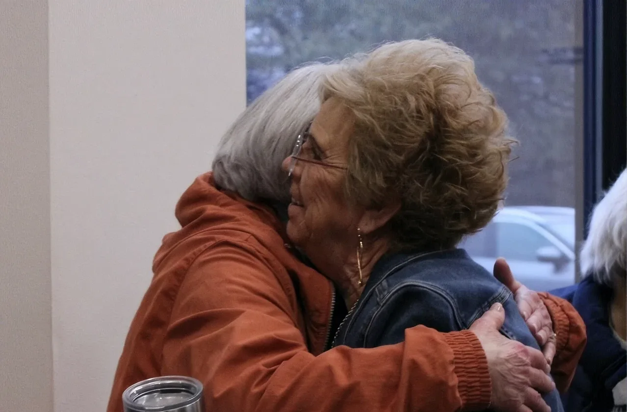 Two women embrace in a warm hug near a window at Journey Church, one holding a stainless steel coffee tumbler.
