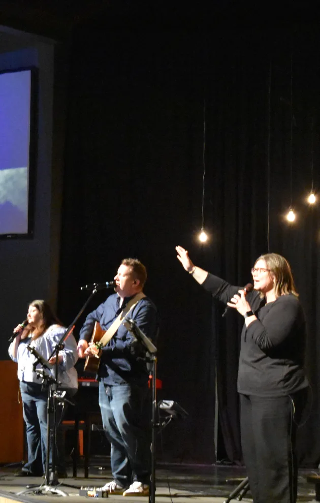 Worship leaders sing and raise a hand during a candlelit moment at a Journey Church service, with the flame projected on the screen behind them.