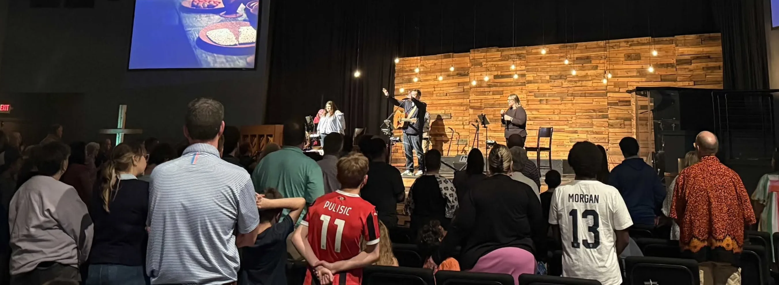 Congregants raise their hands in worship as a band leads music on stage during a Journey Church service.