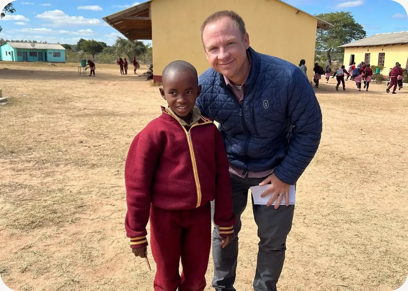 A Journey Church member smiles alongside a child during an international mission trip, with a rural schoolyard in the background.