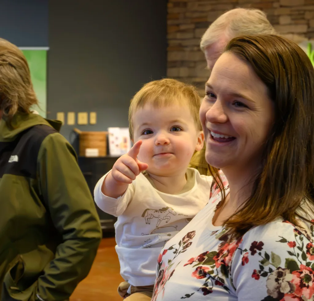 A mom holds a smiling baby who points at the camera while checking in at the Journey Kids welcome desk.