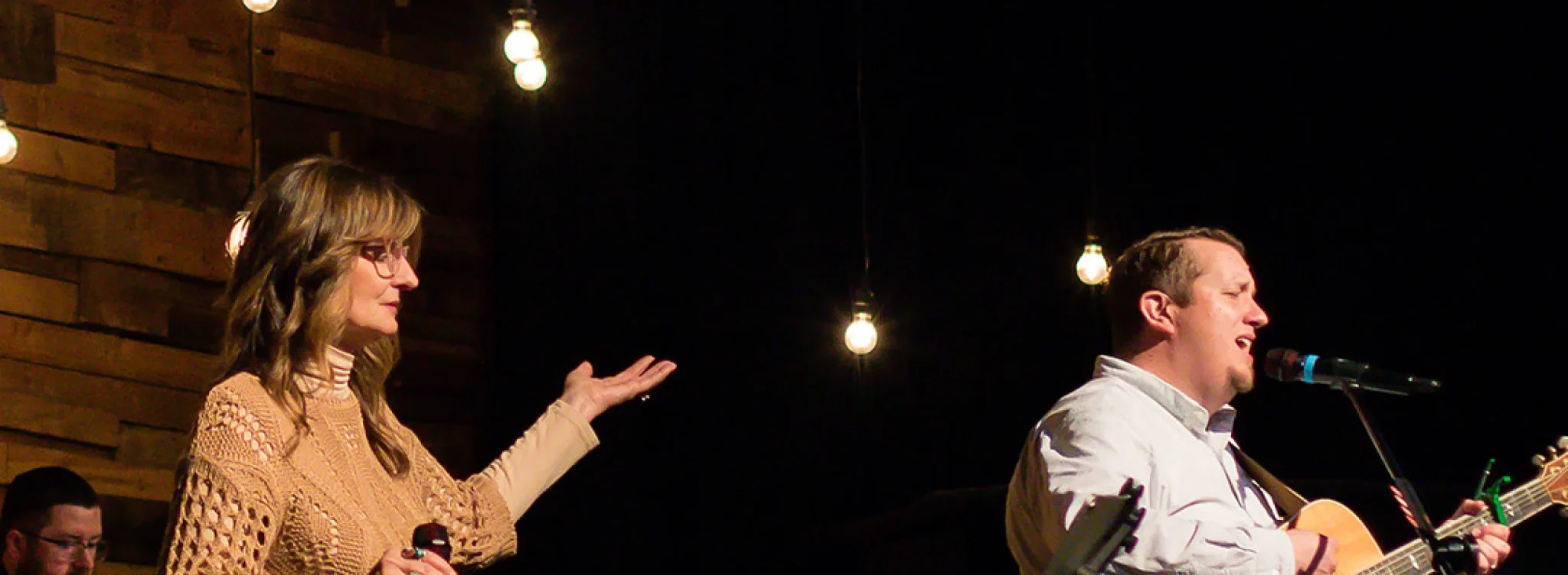 A woman gestures while singing with a mic alongside a man playing acoustic guitar and singing, with string lights behind them on stage.