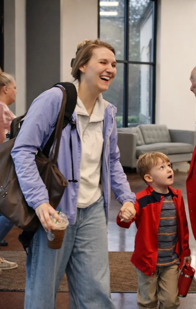 A mom carrying a coffee and backpack smiles while walking hand-in-hand with her young son through a church lobby.