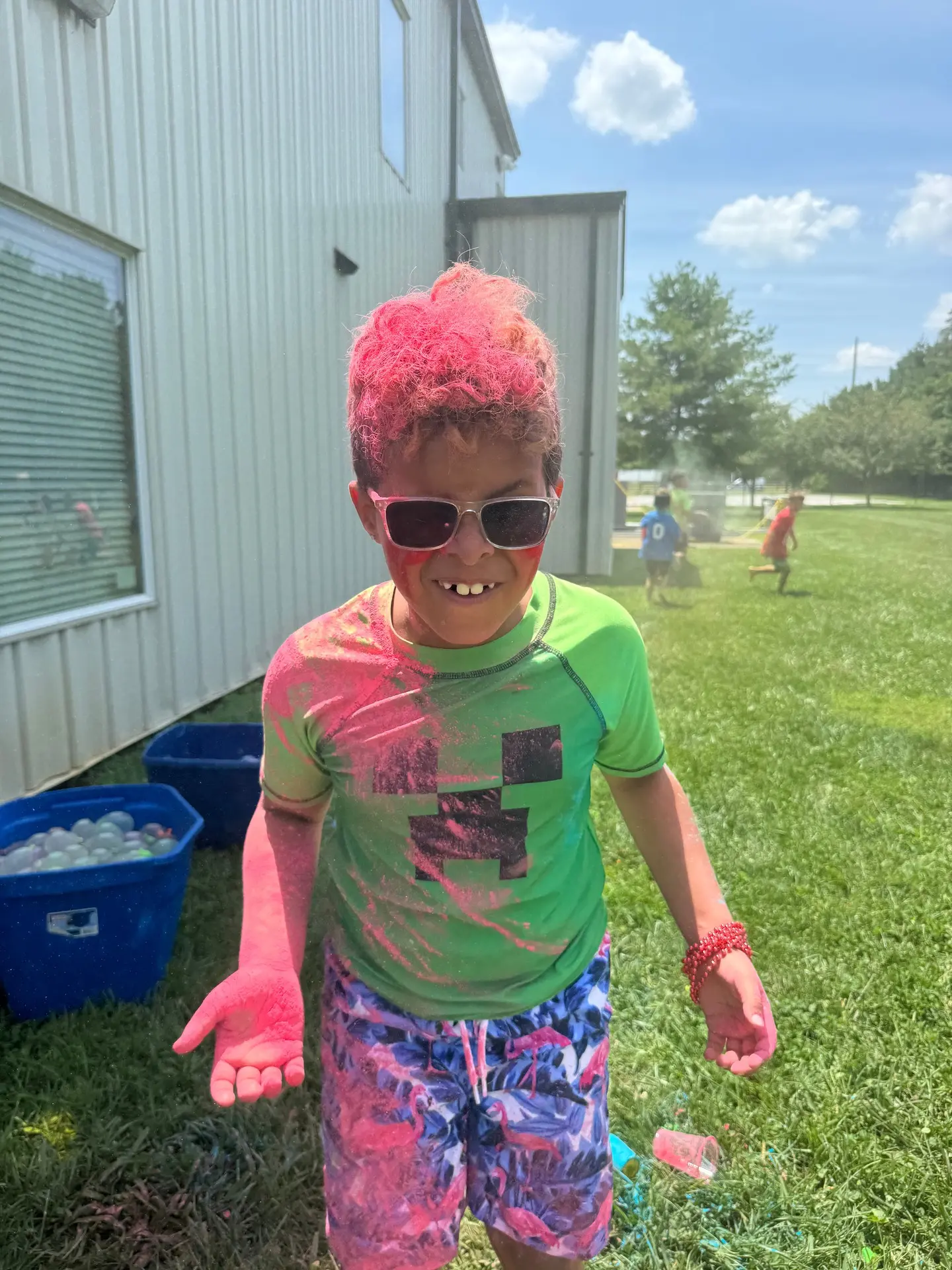 A camper wearing sunglasses and covered in pink and green color powder smiling outside