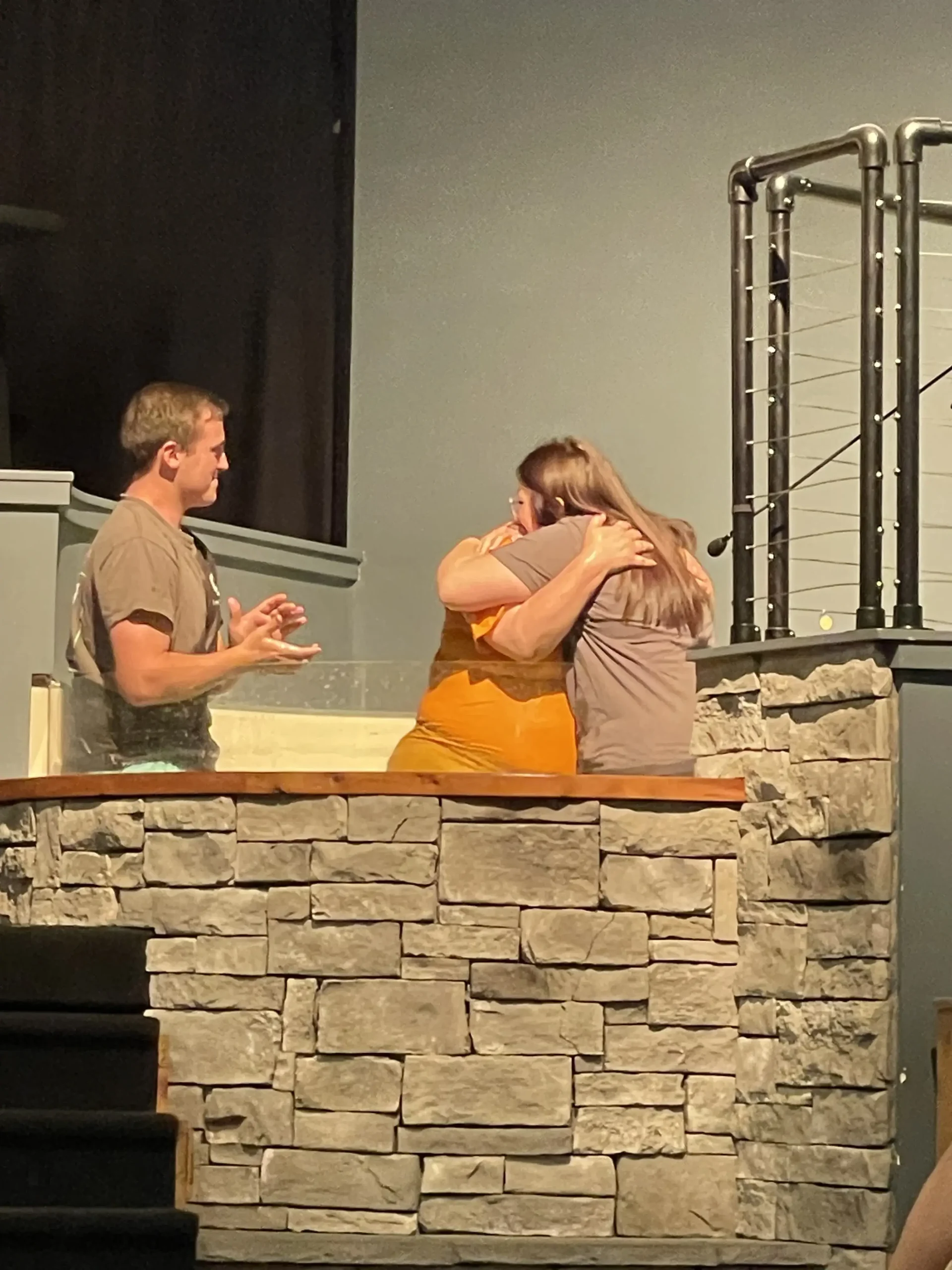 A woman being embraced by a friend in the baptistry after her baptism, with a pastor standing beside them