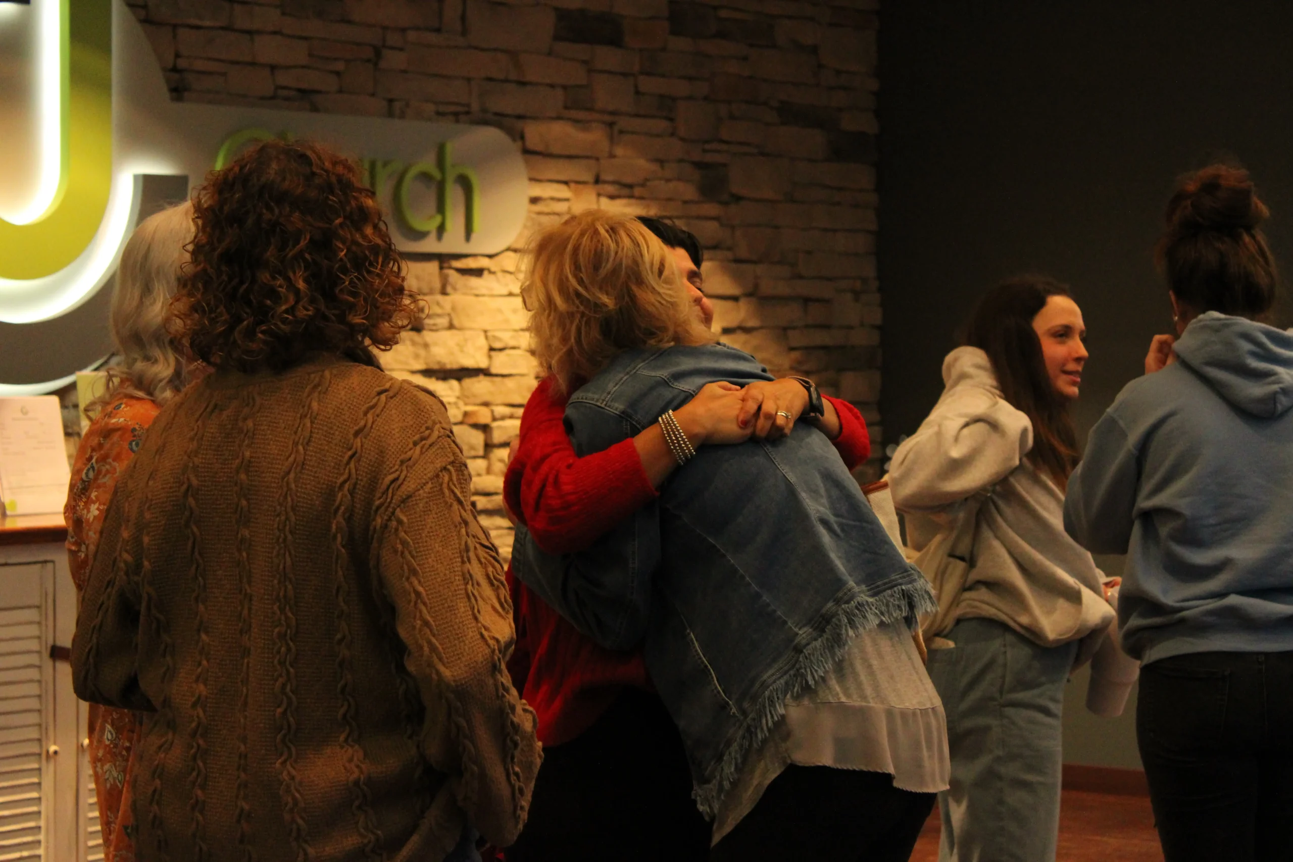 Two women embracing in the Journey Church lobby in front of the logo wall while others talk nearby