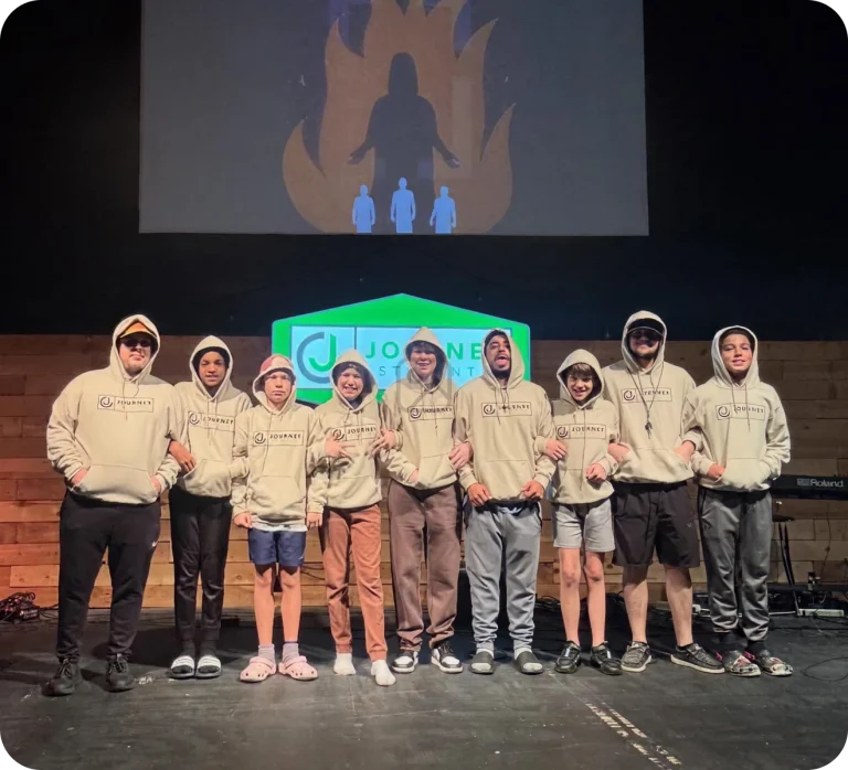 A group of Journey Student teens in matching hoodies pose together on stage in front of the Journey Student sign.