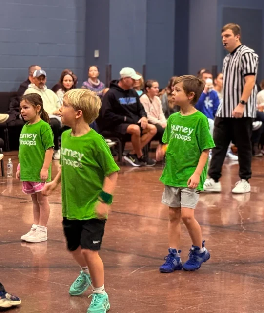 Young players in green Journey Hoops jerseys move down the court during a game while a referee and families watch from the sideline.