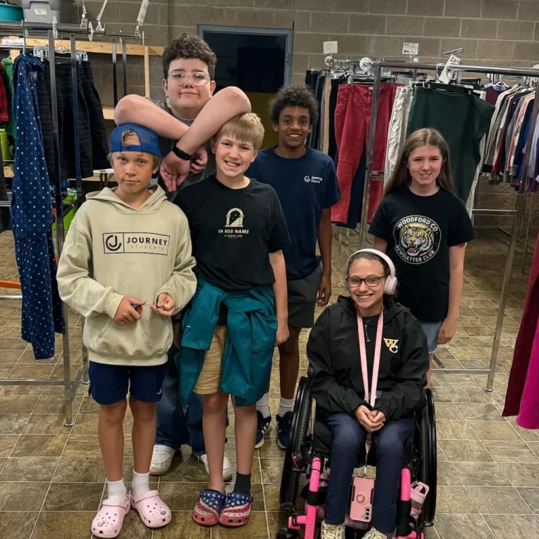 A group of students, including one in a wheelchair, smiles together among the clothing racks while volunteering at Journey Provisions.