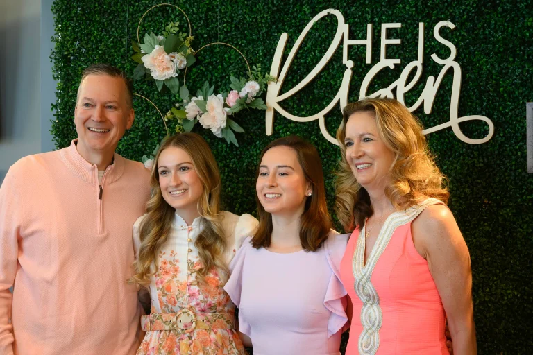 A family of four smiles together in front of a greenery backdrop with a script He Is Risen sign at Journey Church's Easter service.