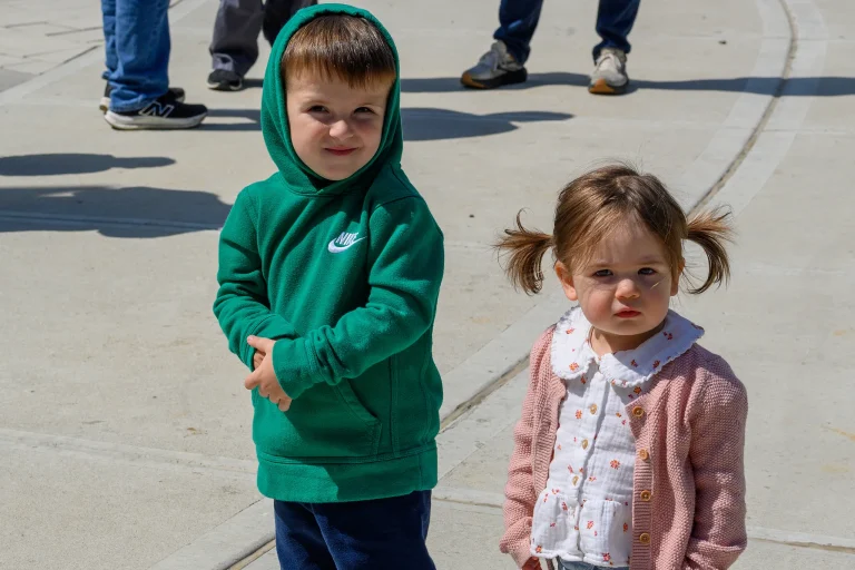 A young boy in a green hoodie and a toddler girl in a pink cardigan stand together on the sidewalk outside Journey Church.