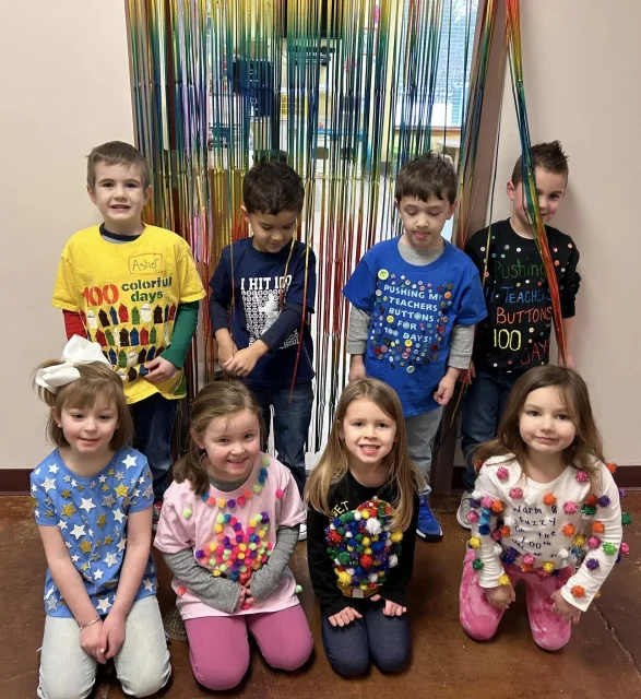 Journey Preschool students pose in two rows in front of a gold fringe backdrop wearing shirts they decorated for the 100th day of school.