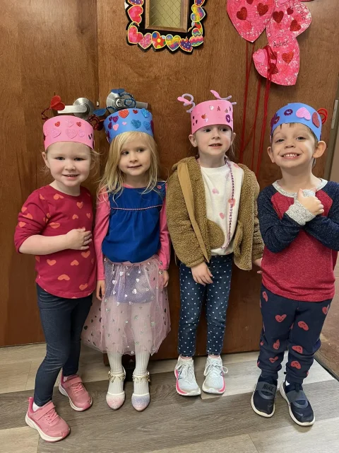 Four Journey Preschool students in Valentine's Day outfits stand together wearing paper crowns they decorated with hearts.