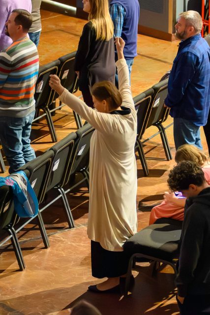 A woman raises one hand in worship among rows of empty chairs during a service.