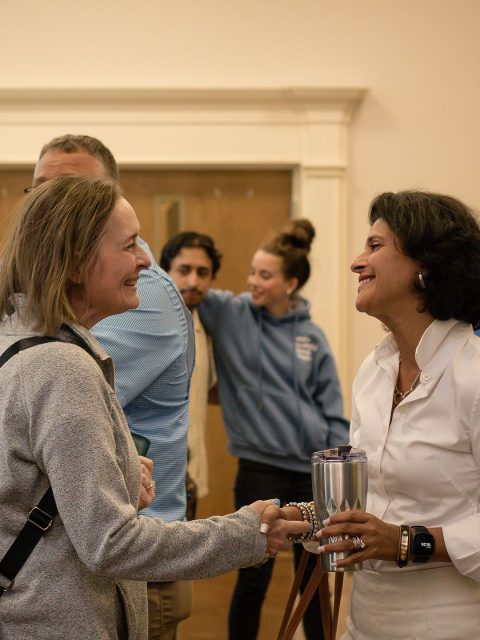 Two women shake hands and smile warmly while greeting each other in the church lobby.