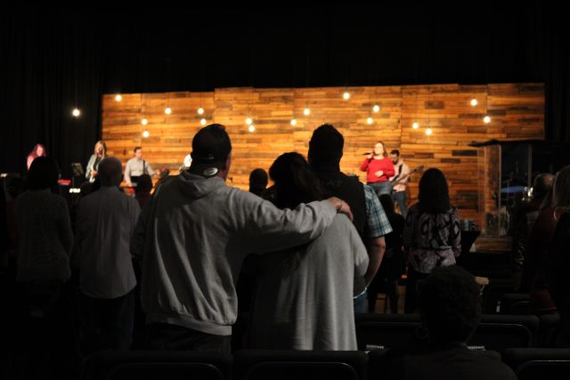 Congregation members stand and worship together, with one couple embracing, as the band plays on stage.