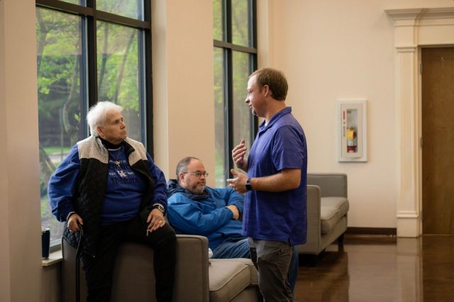 A staff member talks with two seated guests in a lobby seating area.