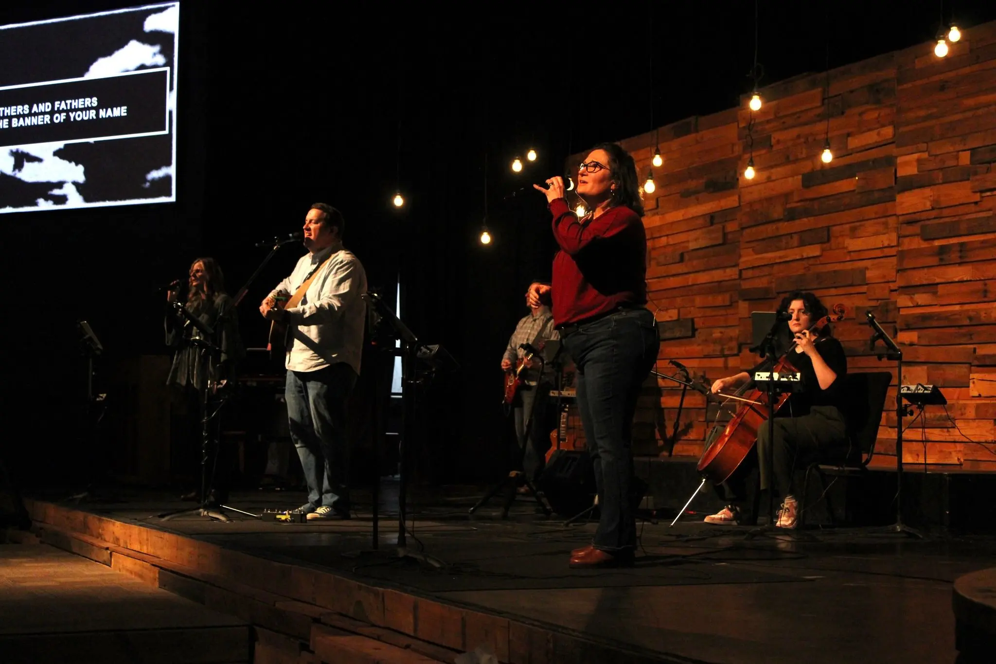 A worship band performs on stage, including guitar, cello, and vocals, under hanging string lights.