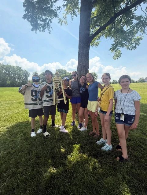A student group posing together under a tree on a sunny lawn during a summer trip