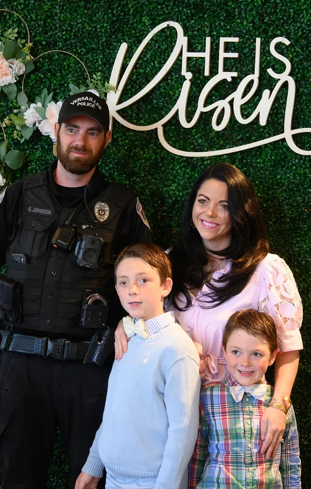 A family, including a dad in a police uniform, poses with their two sons in front of the He Is Risen greenery backdrop at a Journey Church Easter service.