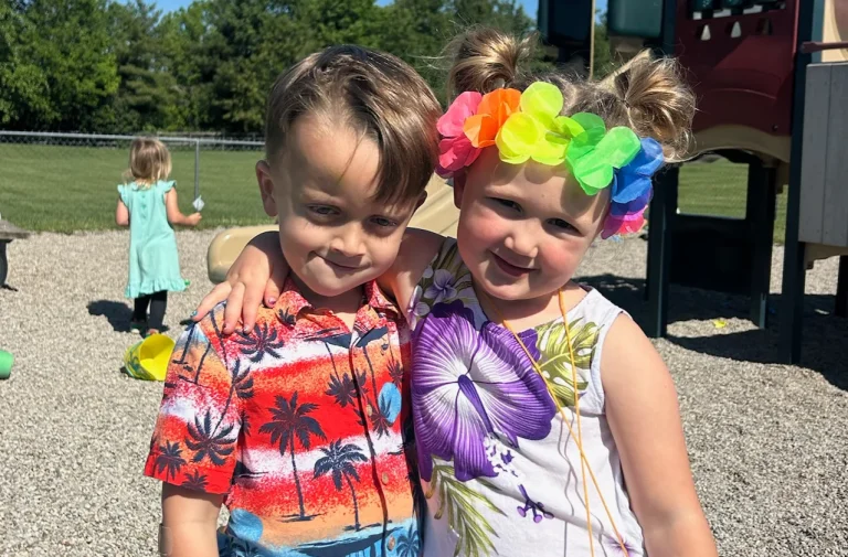 A preschool-age boy and girl in colorful summer outfits smile arm-in-arm on the playground at Journey Preschool.