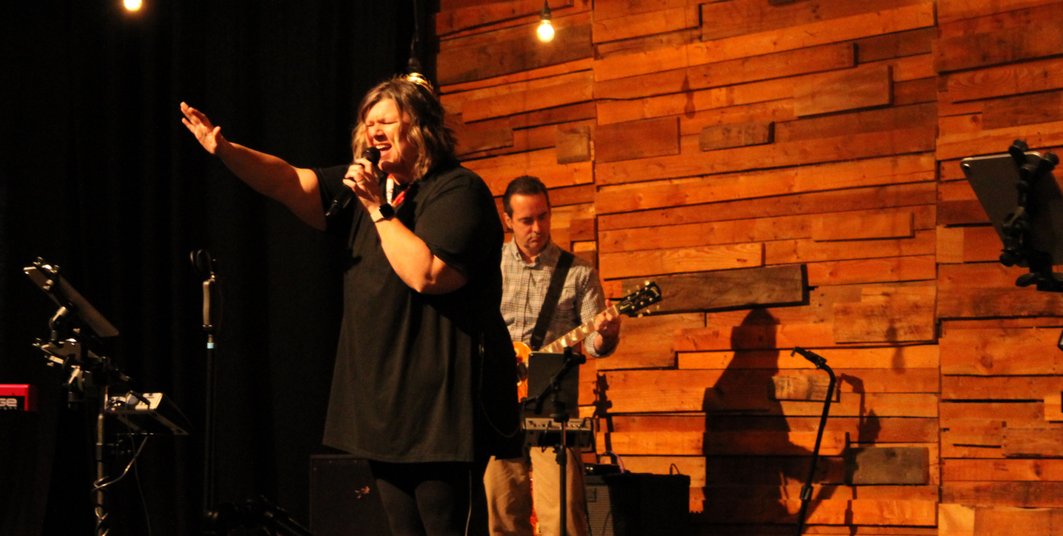 A worship leader sings while raising one hand, silhouetted against a wood-paneled stage.