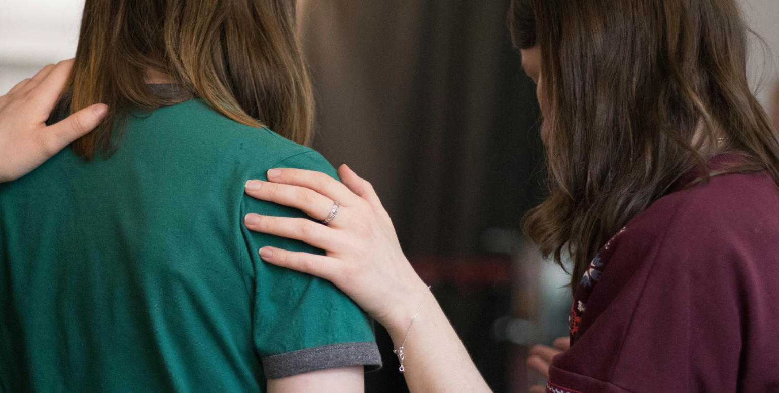Two women stand together from behind, one resting a comforting hand on the other's shoulder.